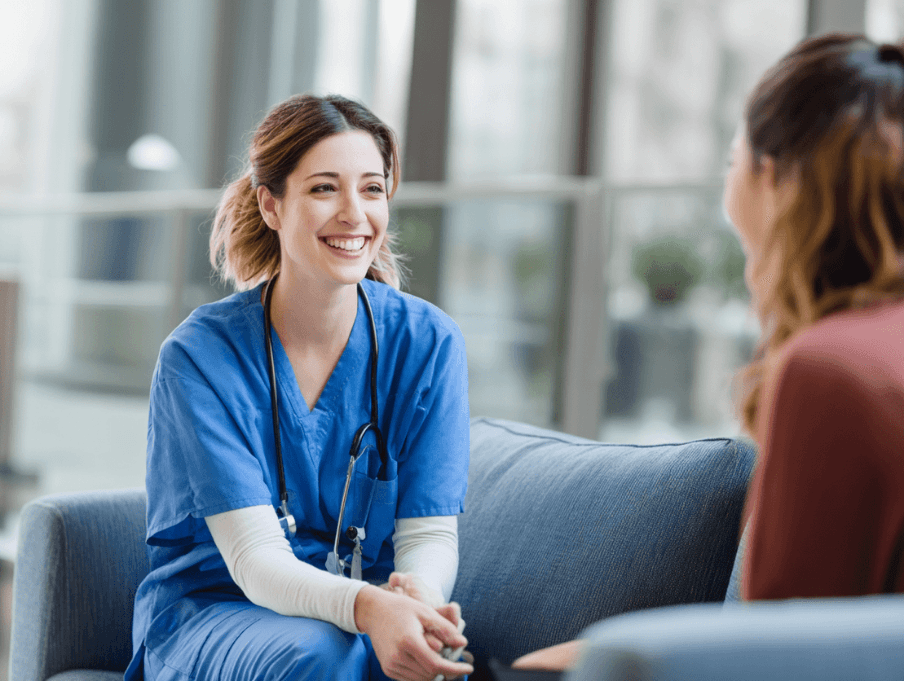 A nurse in blue scrubs with a stethoscope smiling while talking with a patient seated across from her.